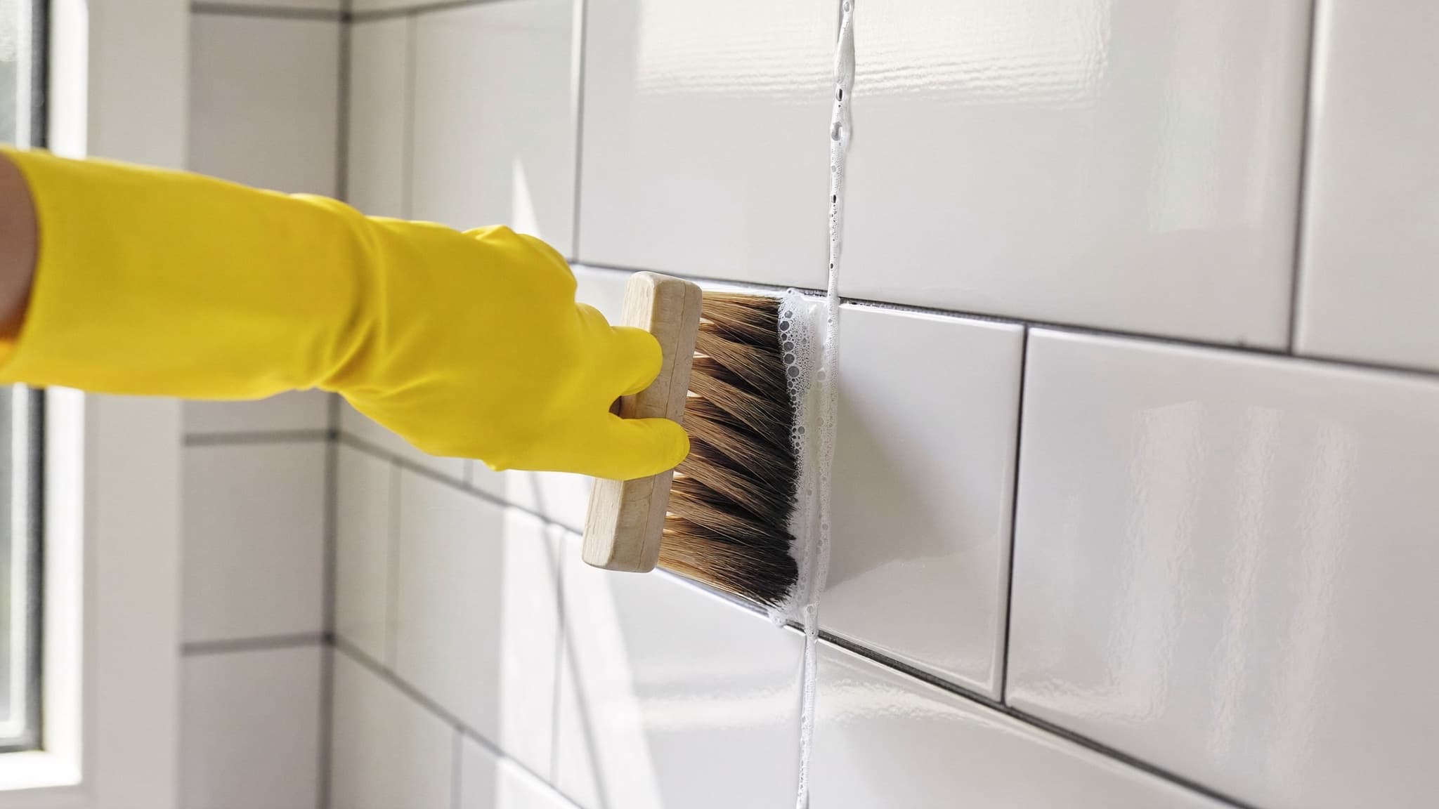 Gloved hand scrubbing white subway tile grout lines with a grout brush in a bright, freshly cleaned bathroom